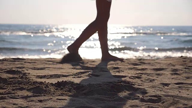 在阳光明媚的日子里，女性的脚在海滩上行走，海浪在背后。年轻女子的双腿踩在沙滩上。暑假或假日概念。侧视图慢动作关闭低角度视图视频素材