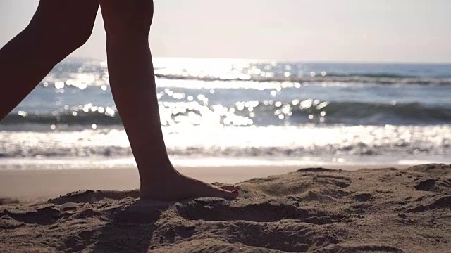 在阳光明媚的日子里，女性的脚在海滩上行走，海浪在背后。年轻女子的双腿踩在沙滩上。暑假或假日概念。侧视图慢动作关闭低角度视图视频素材