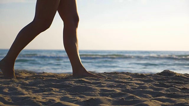 近距离的女性的脚走在海滩与宁静的海浪在背景。年轻女子的双腿踩在沙子上。暑假或假日概念。侧视图慢动作低角度视图视频素材