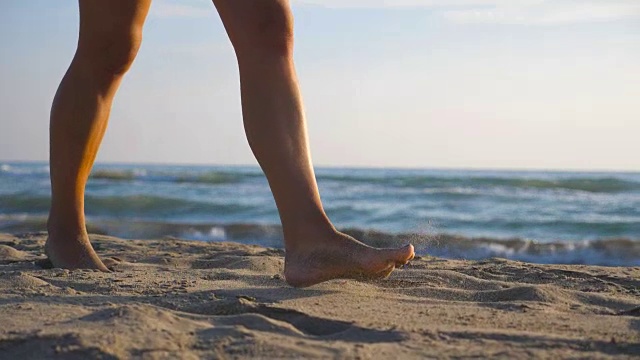 女性的脚踩在沙滩上的特写与海浪的背景。一个美丽的女人在阳光明媚的日子赤脚走在海边。暑假或假期的概念。慢动作低角度视图视频素材