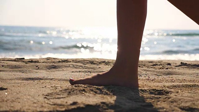 近距离的女性的脚走在金色的沙滩与海浪的背景。年轻女子的双腿踩在沙子上。光着脚的女孩在海边。暑假或假日概念。慢动作低角度视图侧视图视频素材