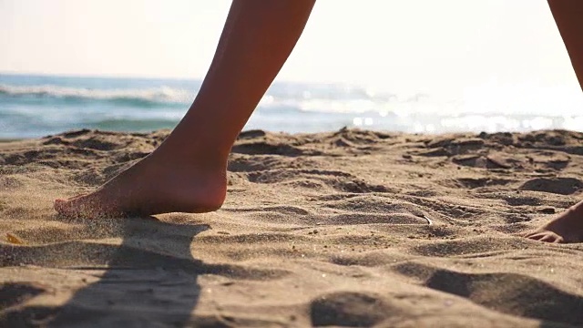 近距离的女性的脚走在金色的沙滩与海浪的背景。年轻女子的双腿踩在沙子上。光着脚的女孩在海边。暑假或假日概念。慢动作低角度视图侧视图视频素材