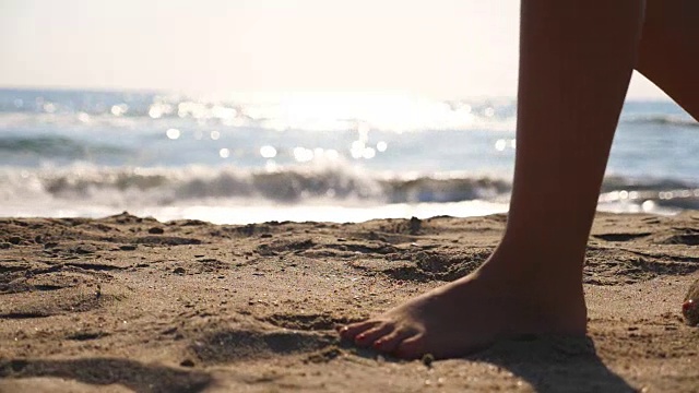 近距离的女性的脚走在金色的沙滩与海浪的背景。年轻女子的双腿踩在沙子上。光着脚的女孩在海边。暑假或假日概念。慢动作低角度视图侧视图视频素材