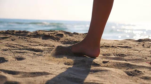 近距离的女性的脚走在金色的沙滩与海浪的背景。年轻女子的双腿踩在沙子上。光着脚的女孩在海边。暑假或假日概念。慢动作低角度视图侧视图视频素材