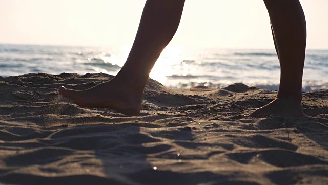 近距离的女性的脚走在金色的沙滩与海浪的背景。年轻女子的双腿踩在沙子上。光着脚的女孩在海边。暑假或假日概念。慢动作低角度视图侧视图视频素材