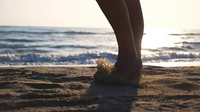 近距离的女性的脚走在金色的沙滩与海浪的背景。年轻女子的双腿踩在沙子上。光着脚的女孩在海边。暑假或假日概念。慢动作低角度视图侧视图视频素材