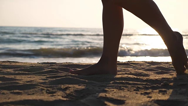 近距离的女性的脚走在金色的沙滩与海浪的背景。年轻女子的双腿踩在沙子上。光着脚的女孩在海边。暑假或假日概念。慢动作低角度视图侧视图视频素材