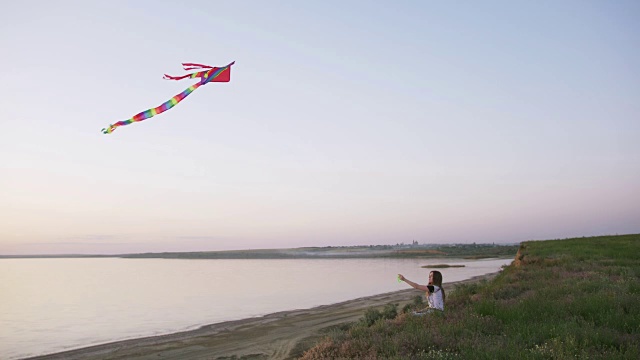 快乐的年轻女子在一个湖泊背景的林间空地上玩风筝在夏季日落视频素材