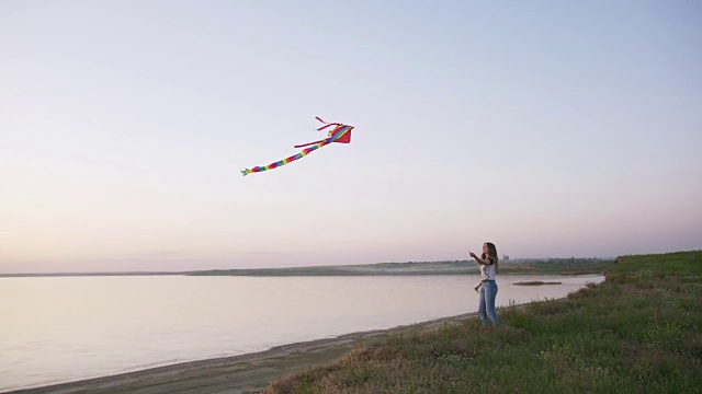 快乐的年轻女子在一个湖泊背景的林间空地上玩风筝在夏季日落视频素材