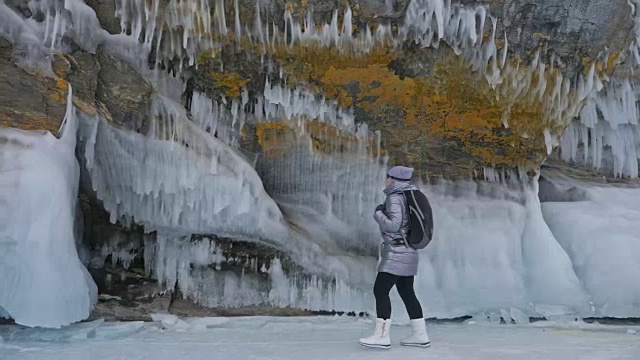 女人在贝加尔湖冰面上的旅行。冬岛之旅。女孩在冰岩石下行走。游客看着美丽的冰洞。极限跋涉和步行。背包客在大自然中休憩。视频素材