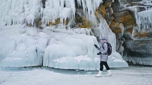 女人在贝加尔湖冰面上的旅行。冬岛之旅。女孩在冰岩石下行走。游客看着美丽的冰洞。极限跋涉和步行。背包客在大自然中休憩。视频素材