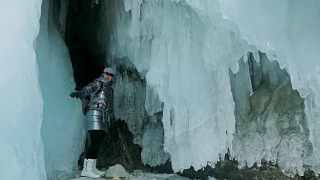 贝加尔湖冰洞上的女游客。冬岛之旅。女孩背包客正在冰洞中行走。游客看着美丽的冰洞。徒步旅行者穿银色夹克，背包，led灯。视频素材