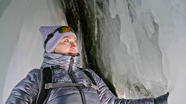 贝加尔湖冰洞上的女游客。冬岛之旅。女孩背包客正在冰洞中行走。游客看着美丽的冰洞。徒步旅行者穿银色夹克，背包，led灯。视频素材