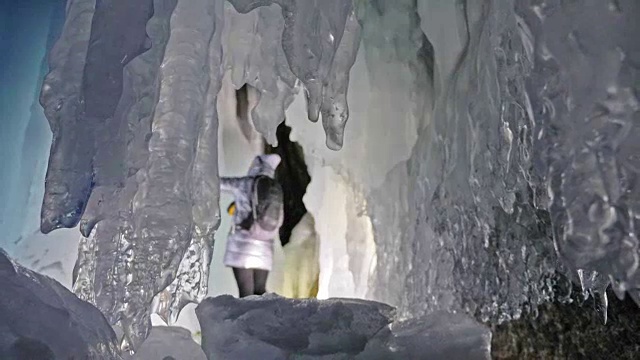 贝加尔湖冰洞上的女游客。冬岛之旅。女孩背包客正在冰洞中行走。游客看着美丽的冰洞。徒步旅行者穿银色夹克，背包，led灯。视频素材