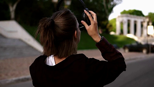 一个年轻的现代女孩在黑色休闲步行通过公园或街道的背影。她回头微笑着。戴上无线耳机。休闲时间视频素材