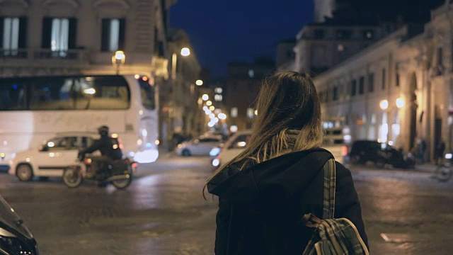 后视图的年轻女游客与背包站在交通道路和环顾在晚上视频素材