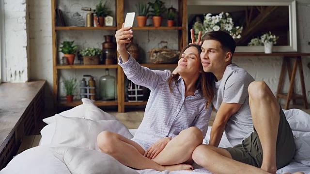 一对快乐恩爱的情侣在家里的床上一起坐着，用智能手机盯着相机自拍，摆姿势，做鬼脸。技术和关系概念。视频素材
