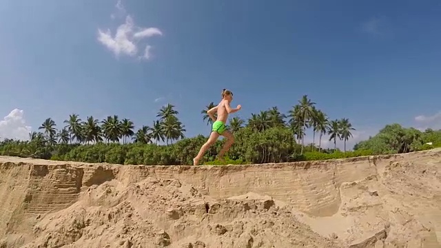 在沙滩上奔跑的小男孩有绿色的棕榈树和蓝天的背景慢镜头视频素材