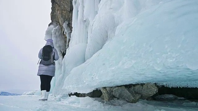 女人在贝加尔湖冰面上的旅行。冬岛之旅。女孩在冰岩石下行走。游客看着美丽的冰洞。极限跋涉和步行。背包客在大自然中休憩。视频素材