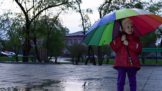 可爱的小女孩拿着五颜六色的雨伞在夕阳下的公园里的水坑里跳跃。视频素材