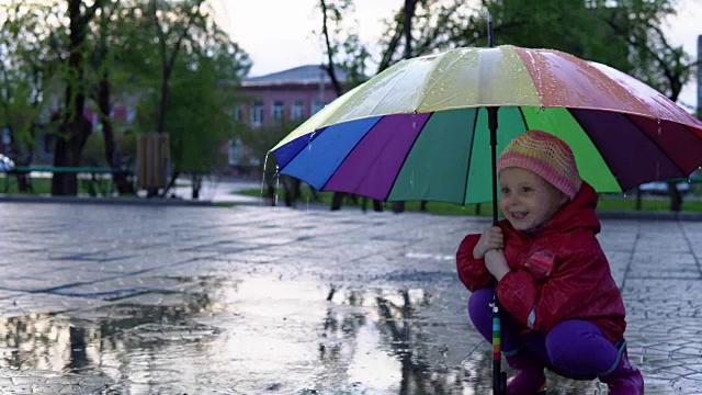 可爱的小女孩拿着五颜六色的雨伞在夕阳下的公园里的水坑里跳跃。视频素材