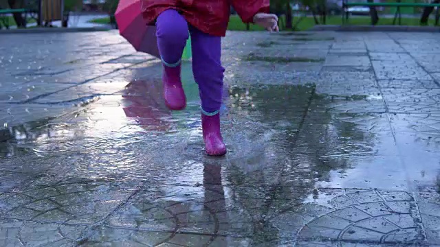 可爱的小女孩拿着五颜六色的雨伞在夕阳下的公园里的水坑里跳跃。视频素材