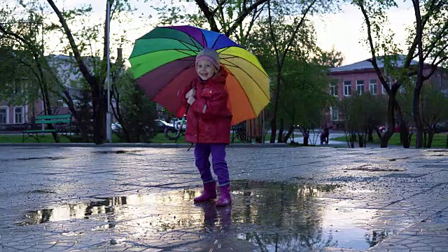 可爱的小女孩拿着五颜六色的雨伞在夕阳下的公园里的水坑里跳跃。视频素材