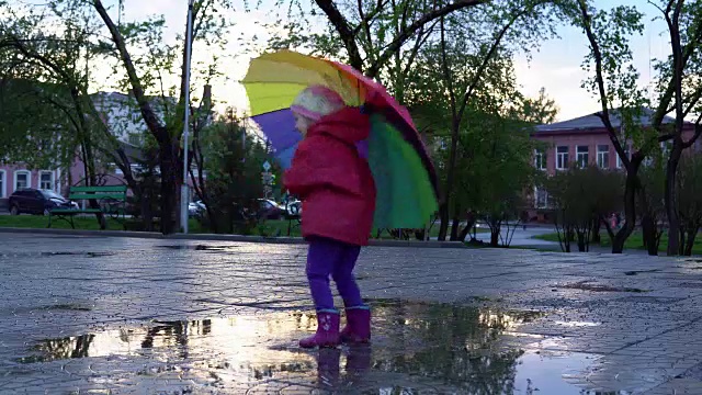 可爱的小女孩拿着五颜六色的雨伞在夕阳下的公园里的水坑里跳跃。视频素材