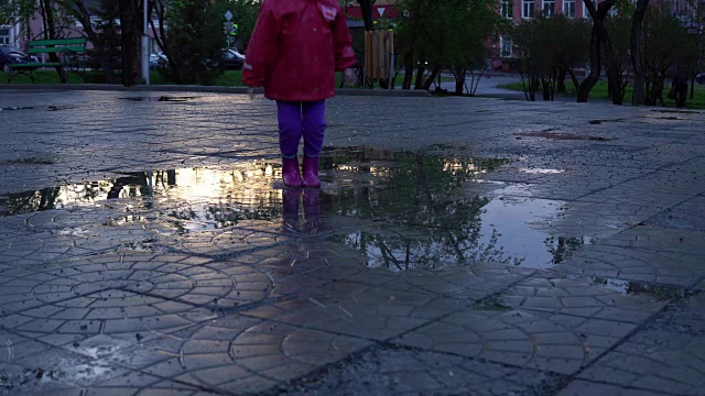 可爱的小女孩拿着五颜六色的雨伞在夕阳下的公园里的水坑里跳跃。视频素材