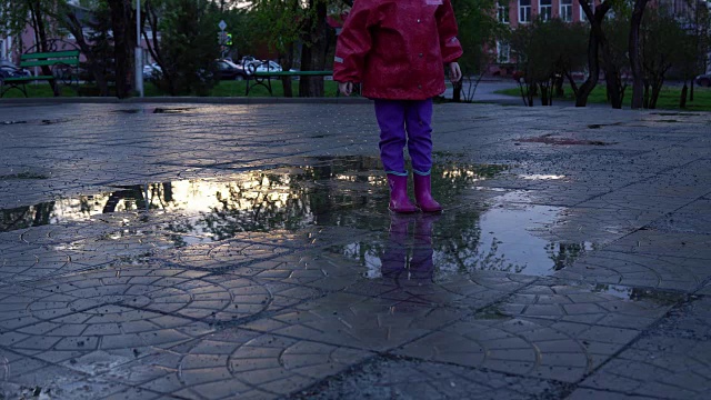 可爱的小女孩拿着五颜六色的雨伞在夕阳下的公园里的水坑里跳跃。视频素材