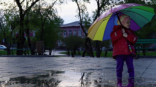 可爱的小女孩拿着五颜六色的雨伞在夕阳下的公园里的水坑里跳跃。视频素材