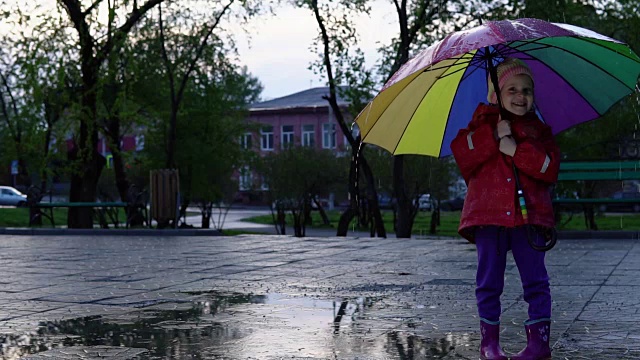 可爱的小女孩拿着五颜六色的雨伞在夕阳下的公园里的水坑里跳跃。视频素材