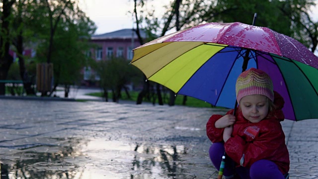 可爱的小女孩拿着五颜六色的雨伞在夕阳下的公园里的水坑里跳跃。视频素材