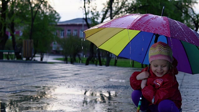 可爱的小女孩拿着五颜六色的雨伞在夕阳下的公园里的水坑里跳跃。视频素材