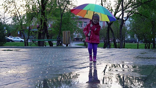 可爱的小女孩拿着五颜六色的雨伞在夕阳下穿过公园的水坑视频素材