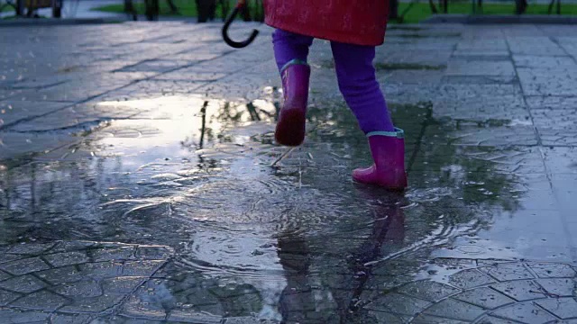 可爱的小女孩拿着五颜六色的雨伞在夕阳下的公园里的水坑里跳跃。视频素材