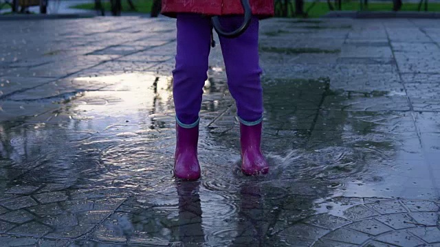 可爱的小女孩拿着五颜六色的雨伞在夕阳下的公园里的水坑里跳跃。视频素材