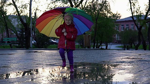 可爱的小女孩拿着五颜六色的雨伞在夕阳下的公园里的水坑里跳跃。视频素材