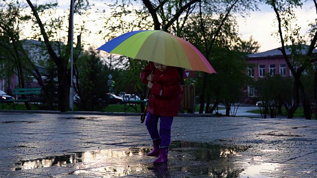 可爱的小女孩拿着五颜六色的雨伞在夕阳下的公园里的水坑里跳跃。视频素材