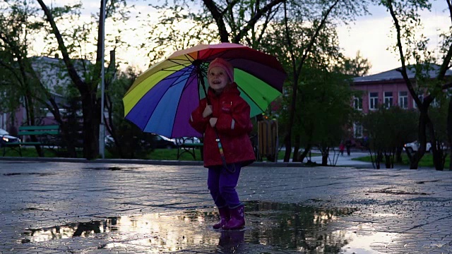 可爱的小女孩拿着五颜六色的雨伞在夕阳下的公园里的水坑里跳跃。视频素材