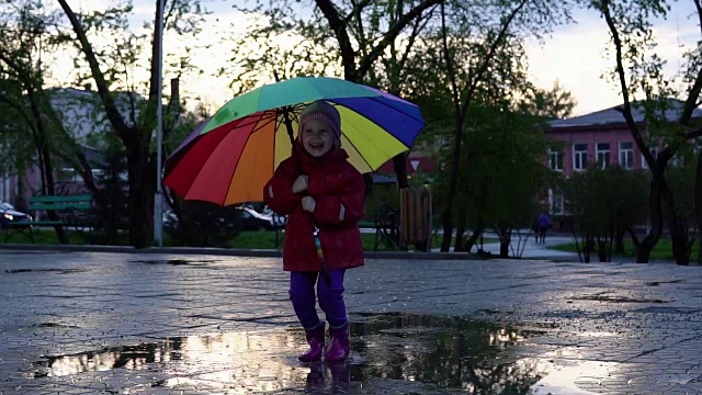 可爱的小女孩拿着五颜六色的雨伞在夕阳下的公园里的水坑里跳跃。视频素材