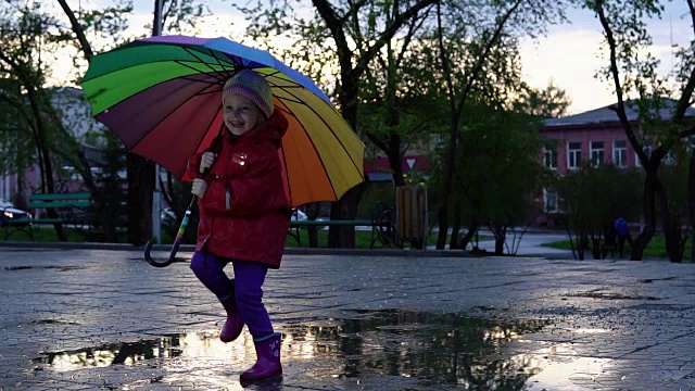 可爱的小女孩拿着五颜六色的雨伞在夕阳下的公园里的水坑里跳跃。视频素材