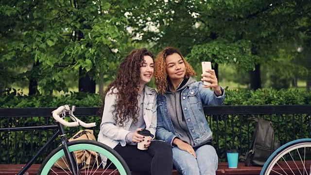 女性朋友拿着咖啡坐在公园的长椅上用智能手机自拍，年轻女性则摆着傻傻的脸享受闲暇时光。视频素材