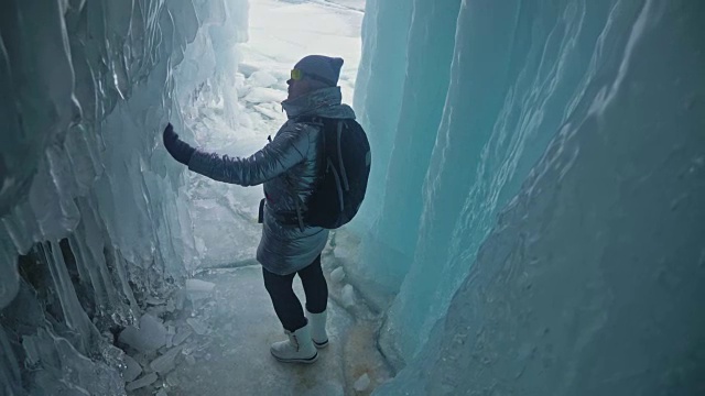 贝加尔湖冰洞上的女游客。冬岛之旅。女孩背包客正在冰洞中行走。游客看着美丽的冰洞。徒步旅行者穿银色夹克，背包，led灯。视频素材