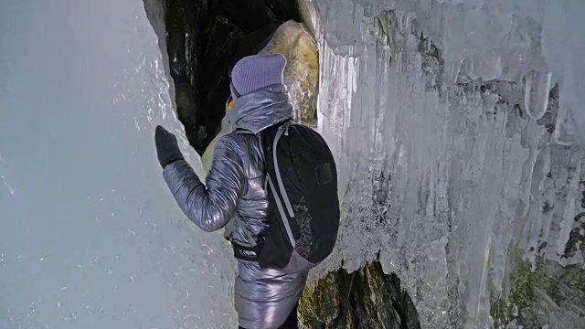 贝加尔湖冰洞上的女游客。冬岛之旅。女孩背包客正在冰洞中行走。游客看着美丽的冰洞。徒步旅行者穿银色夹克，背包，led灯。视频素材