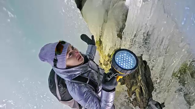 贝加尔湖冰洞上的女游客。冬岛之旅。女孩背包客正在冰洞中行走。游客看着美丽的冰洞。徒步旅行者穿银色夹克，背包，led灯。视频素材