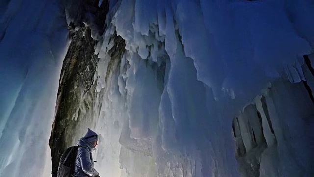 贝加尔湖冰洞上的女游客。冬岛之旅。女孩背包客正在冰洞中行走。游客看着美丽的冰洞。徒步旅行者穿银色夹克，背包，led灯。视频素材