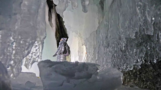 贝加尔湖冰洞上的女游客。冬岛之旅。女孩背包客正在冰洞中行走。游客看着美丽的冰洞。徒步旅行者穿银色夹克，背包，led灯。视频素材