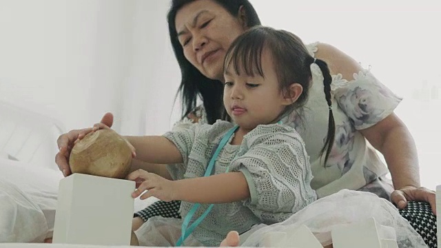 泰国小女孩正在把硬币放进她的白色盒子里，她的母亲和弟弟在她的女儿旁边视频素材