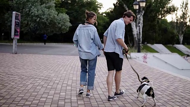 一对年轻的白种人夫妇正牵着狗在城市公园里散步。穿着蓝色衬衫。男孩在散步的时候和他们的狗玩。罕见的视图视频素材